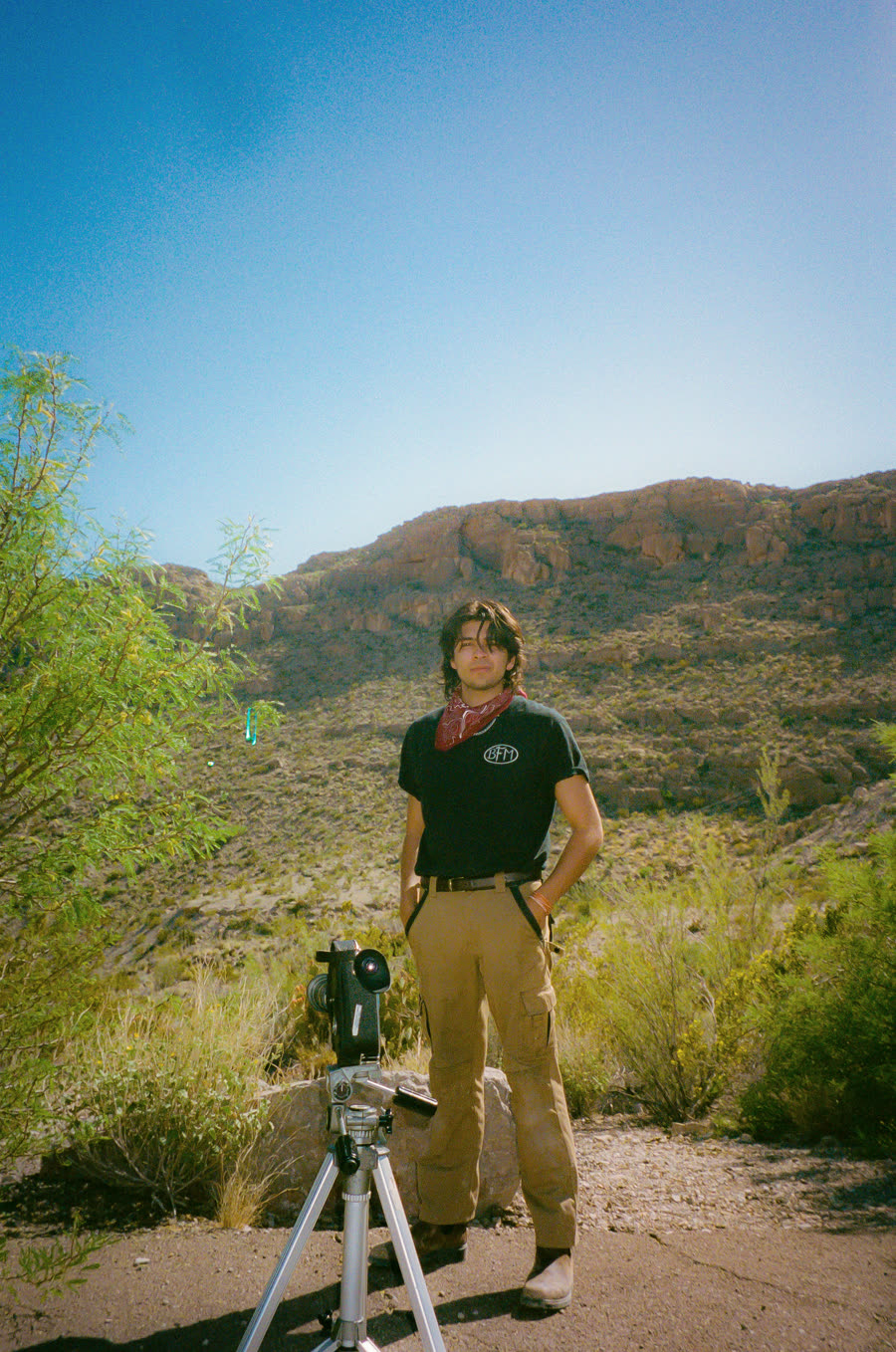 Abraham Thodakee standing with a film camera on a tripod in a desert canyon, red bandana at his neck